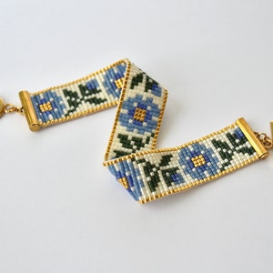 May include: A white beaded bracelet with a blue and green floral pattern. The bracelet has a gold clasp and a gold ring.