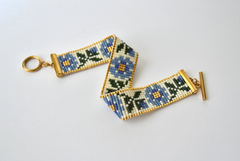 May include: A white beaded bracelet with a blue and green floral pattern. The bracelet has a gold clasp and a gold ring.
