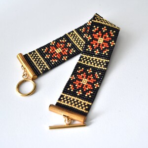 Puede incluir: Un brazalete de cuentas negras, doradas y naranjas con un patrón geométrico. El brazalete tiene un cierre dorado.