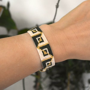 May include: A black and white beaded bracelet with a geometric pattern and gold accents. The bracelet is on a person's wrist.