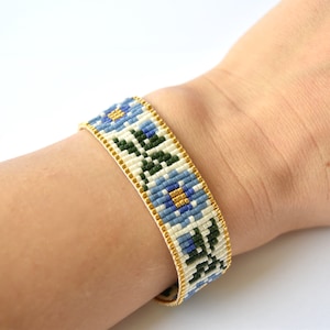 May include: A white beaded bracelet with a floral pattern in blue, green, and gold. The bracelet is on a person's wrist.
