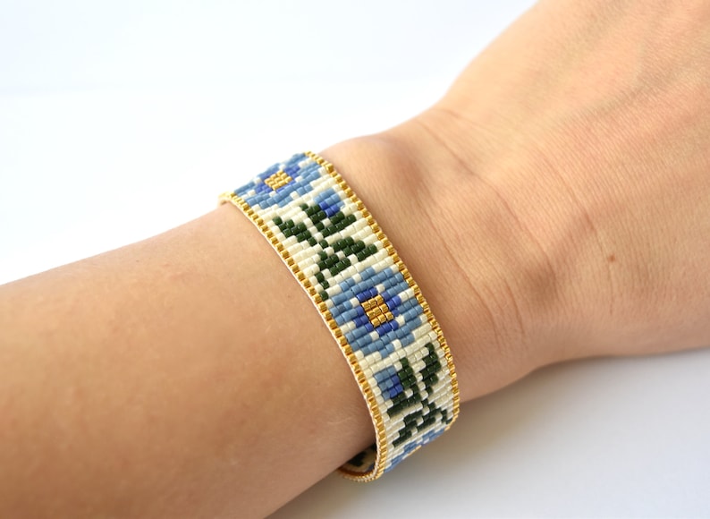 May include: A white beaded bracelet with a floral pattern in blue, green, and gold. The bracelet is on a person's wrist.