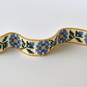 May include: A white beaded bracelet with a floral pattern in blue, green, and gold. The bracelet has a gold clasp and a toggle closure.