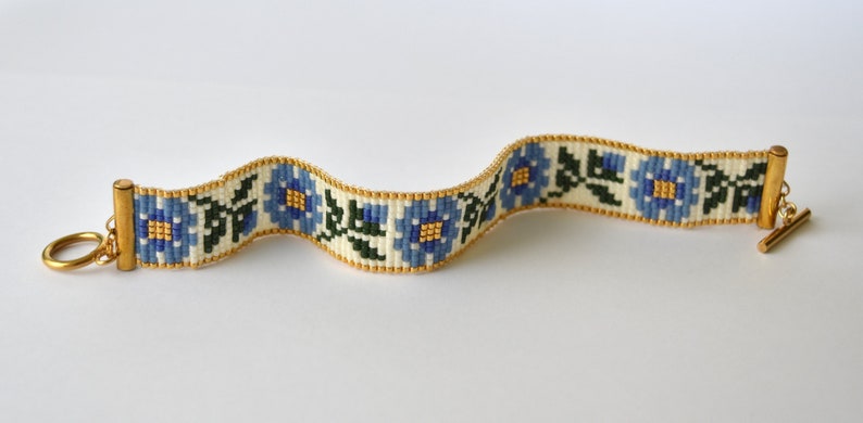 May include: A white beaded bracelet with a floral pattern in blue, green, and gold. The bracelet has a gold clasp and a toggle closure.