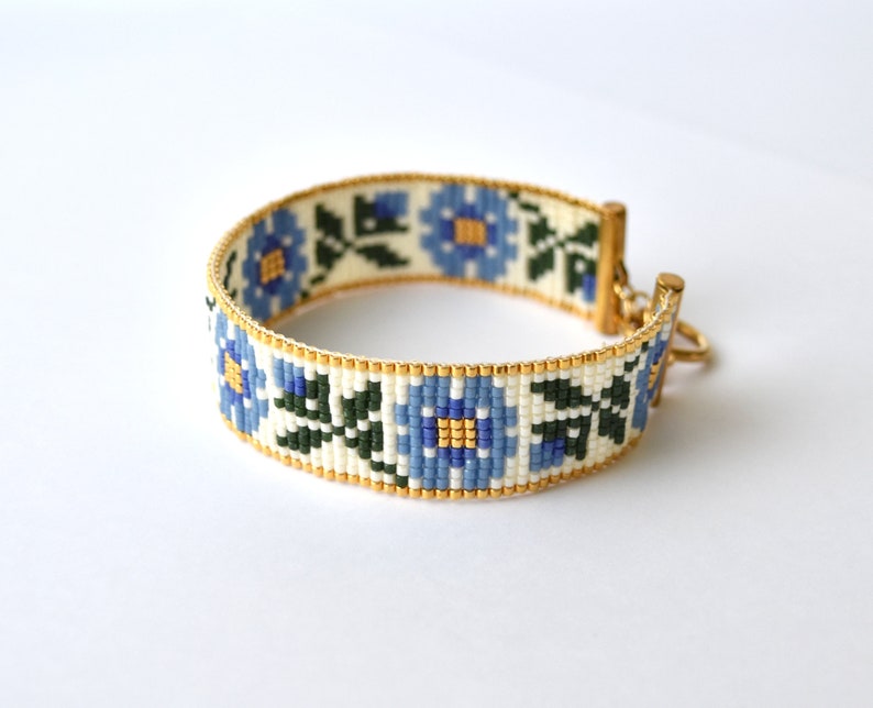May include: A white beaded bracelet with a blue floral pattern and gold accents. The bracelet has a clasp closure.