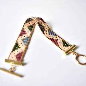 K&ouml;nnte beinhalten: Ein Armband aus Perlen mit einem geometrischen Muster in Rosa-, Gr&uuml;n-, Blau- und Lilat&ouml;nen. Das Armband hat goldfarbene Metallverschl&uuml;sse.