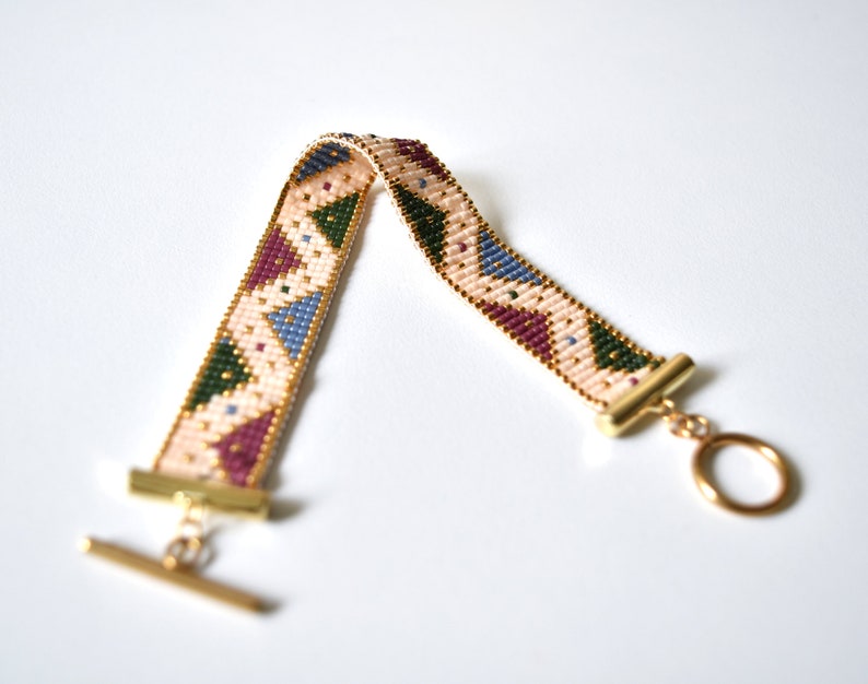 K&ouml;nnte beinhalten: Ein Armband aus Perlen mit einem geometrischen Muster in Rosa-, Gr&uuml;n-, Blau- und Lilat&ouml;nen. Das Armband hat goldfarbene Metallverschl&uuml;sse.