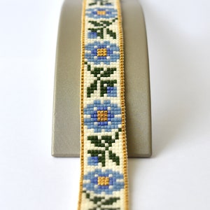 May include: A beaded bracelet with a floral pattern in shades of blue, green, and white. The bracelet is accented with gold trim and a gold clasp.