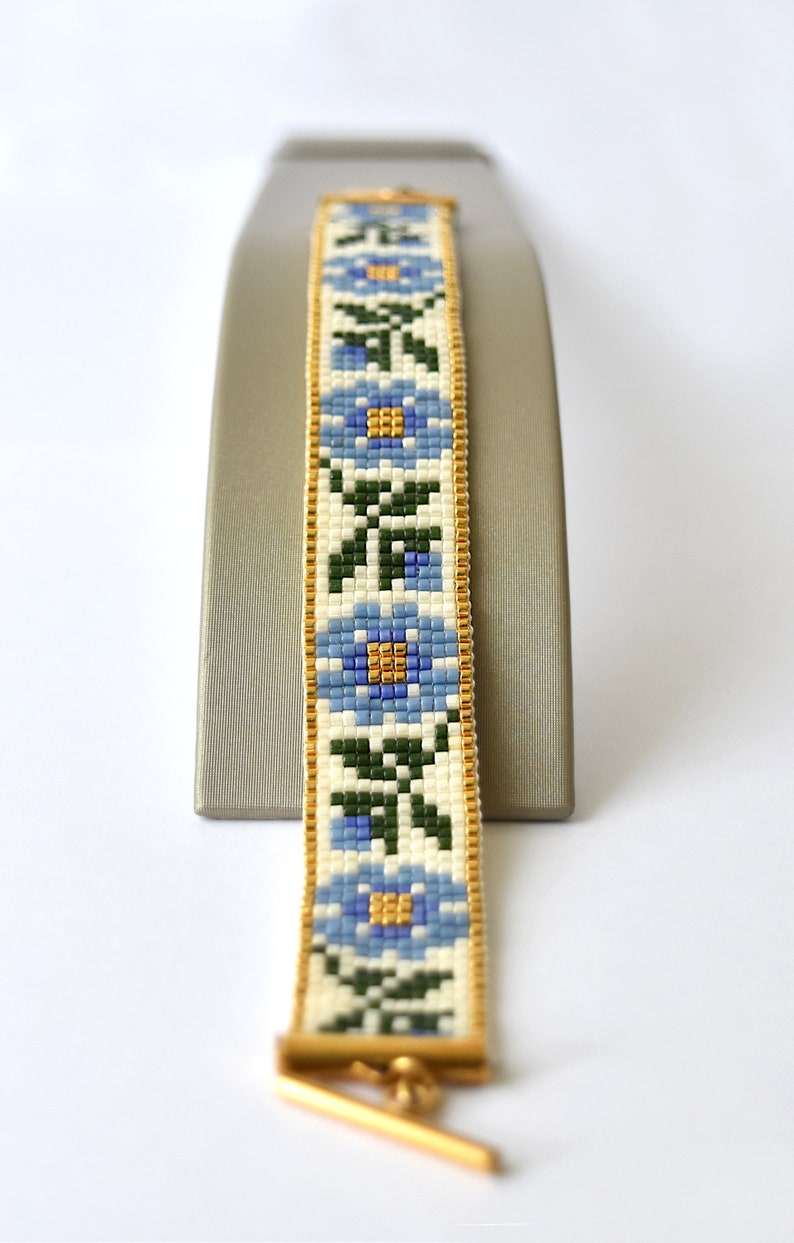 May include: A beaded bracelet with a floral pattern in shades of blue, green, and white. The bracelet is accented with gold trim and a gold clasp.