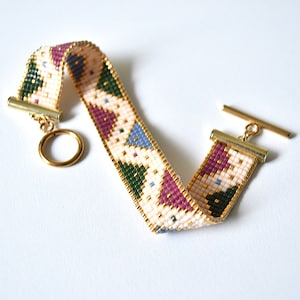 K&ouml;nnte beinhalten: Ein Armband aus Perlen mit einem geometrischen Muster in Rosa-, Gr&uuml;n-, Blau- und Wei&szlig;t&ouml;nen. Das Armband hat einen goldenen Verschluss und einen Schiebeverschluss.