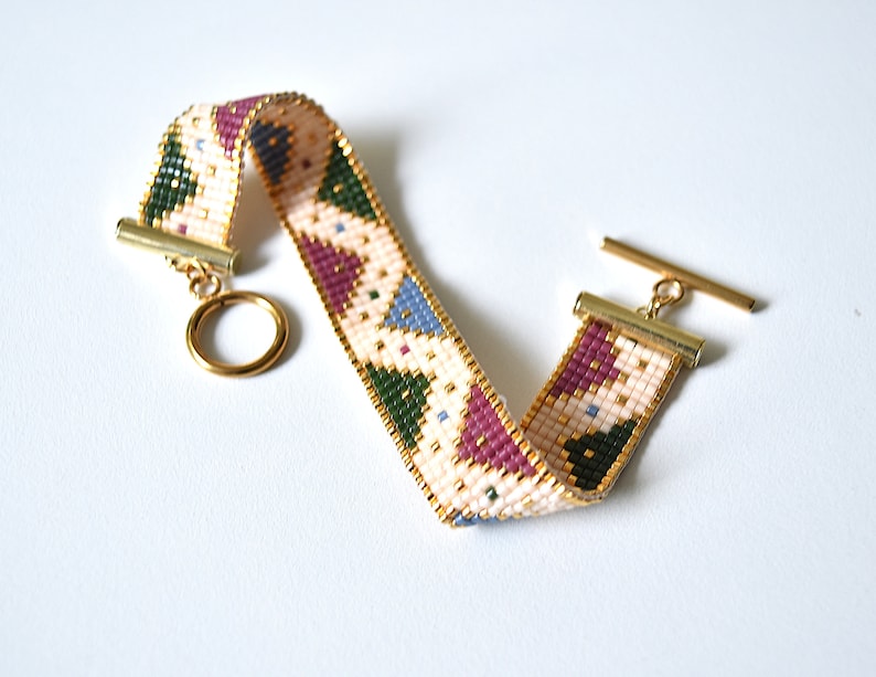 K&ouml;nnte beinhalten: Ein Armband aus Perlen mit einem geometrischen Muster in Rosa-, Gr&uuml;n-, Blau- und Wei&szlig;t&ouml;nen. Das Armband hat einen goldenen Verschluss und einen Schiebeverschluss.
