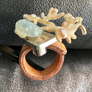 May include: A unique ring featuring a light blue gemstone, a silver square, and coral-like accents with pearls. The ring has a brown leather band, creating a natural and artistic design.
