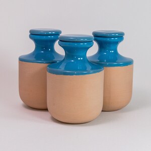 Peut inclure: Trois bocaux de rangement cylindriques avec couvercles assortis. Chaque bocal présente une base en terre cuite lisse non émaillée et une partie supérieure émaillée d'un turquoise brillant et vif, créant un aspect bicolore distinct.