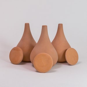 Peut inclure: Trois récipients en terre cuite en forme de goutte d'eau avec des couvercles circulaires assortis. Chaque récipient présente une base large et arrondie qui se rétrécit vers un col étroit. La terre cuite a une finition mate et terreuse.
