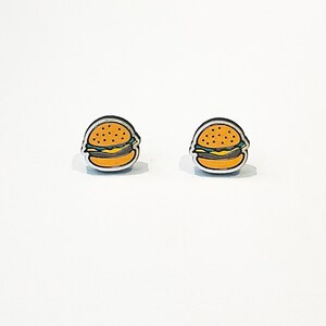 May include: A pair of small, novelty burger-shaped earrings. Each earring features a detailed design with a sesame seed bun, a yellow cheese slice, a green lettuce layer, and a burger patty, all set against a silver border. The earrings are on a white background.
