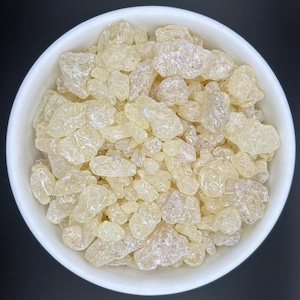 Peut inclure: Un bol en céramique blanc rempli de morceaux de résine jaune pâle, translucides et de forme irrégulière. Les morceaux de résine ont une texture légèrement rugueuse et varient en taille. Le bol est posé sur un fond noir.