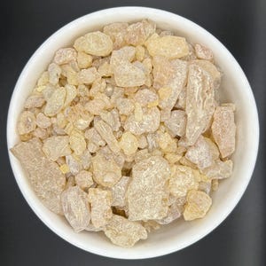 May include: A white ceramic bowl filled with light amber-coloured frankincense resin. The resin pieces vary in size and shape, with a slightly translucent appearance. The bowl is set against a dark background, highlighting the resin's colour and texture.