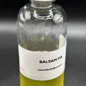 May include: A clear glass bottle with a silver screw cap containing a yellow liquid. A white label on the bottle features the text BALSAM FIR, the website www.blackhillwoods.ca, and the word RESIN printed in red on the side of the label.