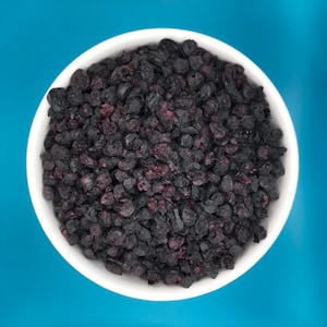 May include: A white bowl filled with dried blueberries. The blueberries are dark purple and have a slightly wrinkled texture.