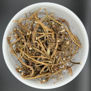 May include: A white ceramic bowl containing dried plant roots. The roots are brown and have a tangled appearance, with visible rootlets and stems. The image is taken from above, highlighting the texture and colour of the herbal product.