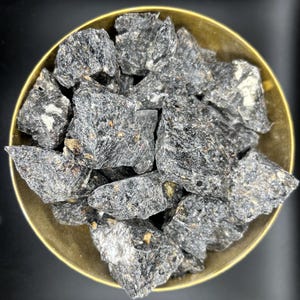 May include: A gold-coloured bowl filled with rough, dark grey stones. The stones have a textured appearance with flecks of lighter colours. The bowl is circular and has a simple, elegant design. The background is dark.