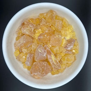 May include: A white ceramic bowl filled with amber-coloured, irregularly shaped resin pieces. The resin chunks vary in size and have a translucent appearance, with some pieces appearing more opaque. The bowl is set against a dark background.