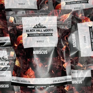 Peut inclure: Plusieurs sachets de thé en feuilles en vrac étiquetés "Black Hill Woods Tea Herbs and Spices" avec la variété "Hibiscus" imprimée sur le devant. Les sachets sont en plastique transparent avec un fond noir et du texte blanc.