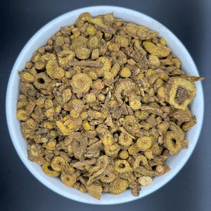 May include: A white bowl filled with dried, chopped, and irregularly shaped pieces of a yellow-brown herb. The pieces vary in size and texture, with some appearing more fibrous and others more solid. The bowl is set against a dark background.