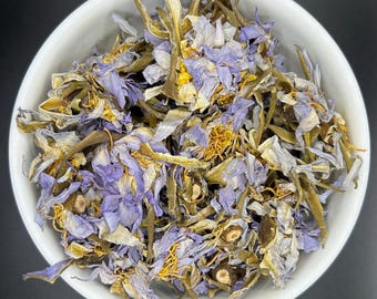 May include: A white bowl filled with dried blue lotus flowers. The mixture consists of light purple and blue petals, thin green stems, and small yellow stamens. The dried botanical elements are densely packed together within the round container.