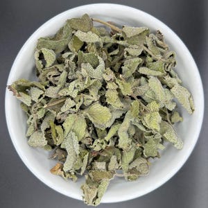 May include: A white ceramic bowl filled with dried sage leaves. The leaves are a muted green color with a slightly fuzzy texture. The sage is a popular herb used in cooking and aromatherapy.