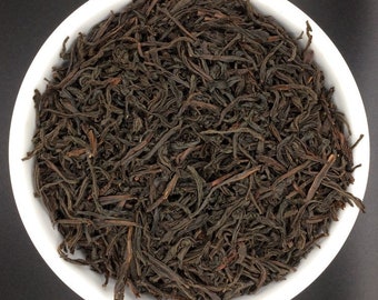 May include: A close-up view of loose black tea leaves in a white bowl. The tea leaves are dark brown and have a slightly reddish hue.
