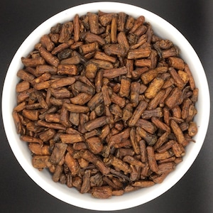 May include: A white bowl filled with dried, brown, stick-shaped pieces of a natural ingredient. The pieces are arranged in a random pattern and appear to be a type of herb or spice.