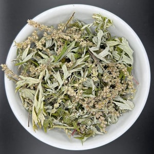 May include: A white ceramic bowl filled with a collection of dried mugwort herbs. The mixture consists of pale green, silvery-grey leaves and small, light brown flower buds, all appearing dehydrated and brittle with a textured, organic appearance.