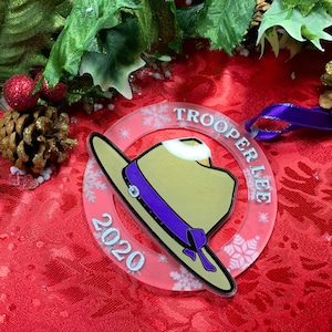 May include: A clear acrylic Christmas ornament with a pink background and white snowflakes. The ornament features a cartoon image of a tan hat with a purple band and a purple bow. The text "TROOPER LEE 2020" is printed in white around the edge of the ornament.
