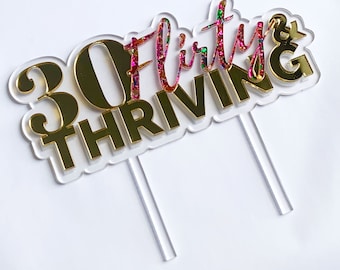 May include: Gold acrylic cake topper with the text "30 & Flirty & Thriving" in a pink glitter font. The number 30 is in a gold font.