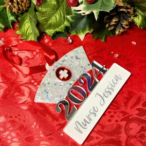 May include: A silver glitter nurse hat Christmas ornament with a red cross and the year 2024. A white rectangular tag is attached to the ornament with the text "Nurse Jessica".