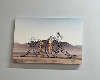 Op de afbeelding: Een zwart metalen sculptuur van twee figuren, een zittend en de ander staand, in een woestijnlandschap. De figuren zijn gemaakt van draad en zijn hol, met zichtbaar inwendige. Het beeldhouwwerk is een hedendaags kunstwerk.