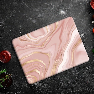 Può includere: Un tagliere in marmo rosa e oro con sfondo bianco. Il tagliere è su una superficie nera con vari alimenti intorno.