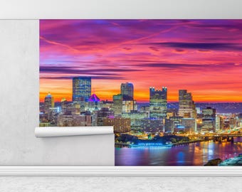May include: A panoramic view of the Pittsburgh skyline at sunset. The city is illuminated with lights, and the sky is a vibrant mix of pink, orange, and purple. The river in the foreground reflects the city lights.