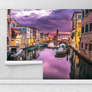 May include: A colorful sunset over a canal in Venice, Italy.  The canal is lined with buildings and there are boats docked along the sides.  A bridge crosses the canal in the middle of the image.