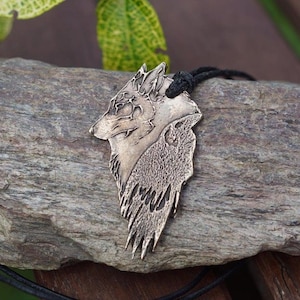Può includere: Una collana con un ciondolo a forma di testa di lupo in argento su un cordoncino nero. Il ciondolo è dettagliato con una superficie testurizzata e un design a forma di testa di lupo.
