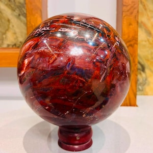 Op de afbeelding: Een gepolijste, bolvormige stenen orb met dieprode, bruine en zwarte wervelende patronen en een glanzend, reflecterend oppervlak. De bol is gemonteerd op een kleine, ronde, donkerrode houten standaard die een stabiele basis voor de display vormt.