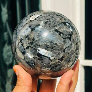 Könnte beinhalten: Eine polierte Kugel mit einem gesprenkelten Muster aus Grau, Schwarz und Silber. Die Kugel wird in einer Hand gehalten, wodurch ihre glatte, reflektierende Oberfläche hervorgehoben wird. Das Design deutet darauf hin, dass es sich um einen Dekorations- oder Sammlerartikel handelt.