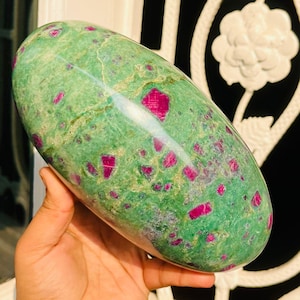 May include: A polished, oval-shaped stone featuring a mottled green base color with vibrant red inclusions. The stone's surface is smooth and reflective, showcasing the natural patterns of the mineral. The stone is held in a hand.