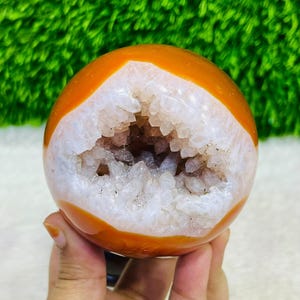 Op de afbeelding: Een bolvormige geode met een gladde, gepolijste oranje buitenkant en een grote, onregelmatige opening die een cluster van doorschijnende, grillige witte kwartskristallen aan de binnenkant onthult. De steen heeft een ronde vorm en glans.