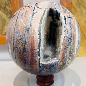 Puede incluir: Una esfera decorativa de piedra con una gran cavidad. La piedra presenta una mezcla de colores beige, rojo y azul, con un interior cristalino. Se exhibe sobre un pequeño soporte redondo de madera oscura.