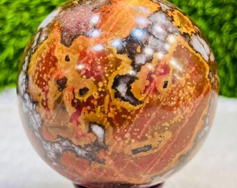 May include: A polished gemstone sphere with a complex pattern of red, brown, orange, and white hues. The sphere is mounted on a dark wooden stand, showcasing its intricate natural design. The sphere's surface has a marbled appearance.