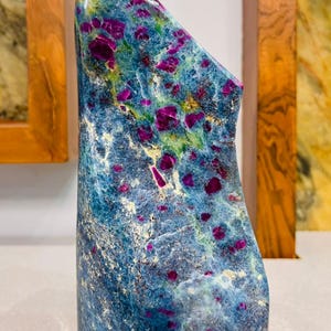 Peut inclure: Une sculpture abstraite et haute en pierre polie. La pierre est principalement bleue avec des stries vertes et jaunes, et de grandes inclusions magenta vibrantes. La sculpture a une base lisse et arrondie et un sommet incliné.
