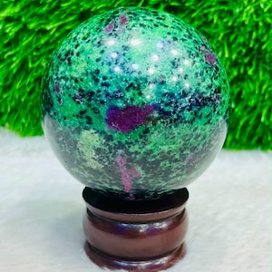 Può includere: Una pietra sferica lucida caratterizzata da una base verde vibrante con inclusioni nere punteggiate e prominenti macchie color magenta intenso. La sfera è esposta su un supporto circolare in legno marrone scuro con finitura liscia.
