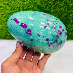 Può includere: Una pietra levigata di forma ovale caratterizzata da una base liscia di colore verde chiaro con inclusioni cristalline irregolari di colore magenta intenso sparse sulla superficie. La pietra presenta una finitura lucida e riflettente.